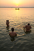 Three devotees wash in the Ganges river waters while a boat passes and the sun is rising in the background