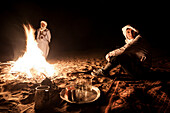 A young woman with turban sits on a carpet over the sand of the desert at night, with a berber man in the background while the fire burns, Sahara desert, Morocco