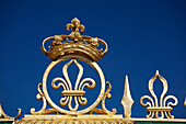 'Gold Crown And Fleur-De-Lis On The Grand Trianon Gates Against A Blue Sky In The Gardens Of The Palace Of Versailles; Paris, France'