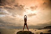 'A Woman Tourist Wearing A Bikini Raises Her Arms And Does Yoga On The Beach Of A Tropical Island At Sunset; Koh Lanta, Thailand'