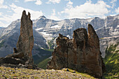 'Large Rock Sentinels On Mountain Ridge With A Mountain Range In The Distance With Blue Sky And Clouds; Alberta, Canada'