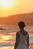 'A young woman stands at the water's edge watching the sunset;Aphrodite bay, cyprus'
