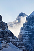 'Sunlit snow blowing off a mountain peak in a v shaped rock cliff with blue sky;Lake louise alberta canada'