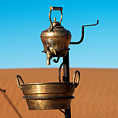 A metal teapot with a tap and bucket below with a sandy landscape and blue sky