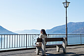 'A Woman Sitting On A Bench Overlooking Lake Maggiore; Brissago, Ticino, Switzerland'
