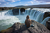 Woman sitting on a rock above Godafoss waterfall, Nordurland Eystra, Iceland