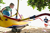 Junge klettert auf traditionellem Fischerboot, 3 Jahre alt, am Strand, traditionelle Boote, prahu, bemalt, Auge, Elternzeit, Elternzeitreise, spielen, Sonnenhut, Meer, Indischer Ozean, MR, Sanur, Bali, Indonesien