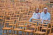 A couple wearing rain capes sitting on their own amidst wooden chairs in the pouring rain outside Friedrichstein Palace, Bad Wildungen, Hesse, Germany, Europe