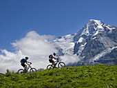 Two mountain bikers are riding through the grassy fields of the Murren plateau. In the background the famous Moench mountain.
