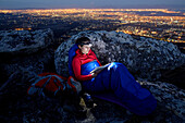 Susann Scheller is sitting against a rock in her sleeping bag while reading a book with a headlamp. In the back down below the lights of the city of Cape Town are shimmering. Table Mountain National Park. South Africa.