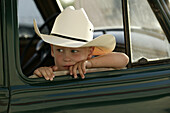 A little boy wearing a cowboy hat leans on the window frame of an old green pick-up truck.