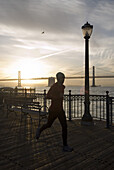 A man running on a dock in the harbour of San Francisco at sunrise with the Oakland Bay Bridge in the background. California, USA.