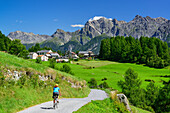 Woman cycling towards Bos-cha, Sesvenna Alps in background, Lower Engadin, Canton of Graubuenden, Switzerland