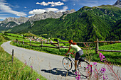 Woman cycling towards Ardez, Sesvenna Alps in background, Lower Engadin, Canton of Graubuenden, Switzerland