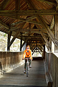Cyclist passing roofed wooden bridge above Inn river, Urgen, Fliess, Tyrol, Austria