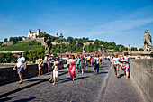 Tourists crossing the Alte Mainbruecke bridge across the Main river with Marienberg fortress on the hillside, Wuerzburg, Franconia, Bavaria, Germany