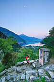 Woman hiking standing in front of chapel, Lago di Mezzola in background, Val Codera, Sentiero Roma, Bergell range, Lombardy, Italy
