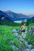 Woman hiking on flagged track through meadow, Lago di Mezzola in background, Val Codera, Sentiero Roma, Bergell range, Lombardy, Italy