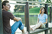 Couple sitting apart on dock, focus on man in foreground