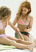 Girls sitting on beach, one applying sunscreen to the other