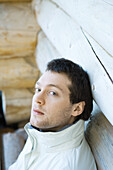 Young man leaning against wall of log cabin, looking at camera