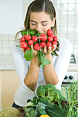 Young woman in kitchen with an assortment of vegetables, smelling bunch of radishes