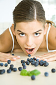 Fresh blueberries scattered across counter, young woman opening mouth to eat one