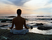 Man sitting in lotus position in front of sea, rear view, silhouette