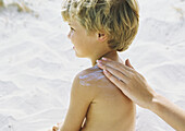 Little boy on beach having sunscreen rubbed into shoulder by mother
