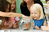 Elementary teacher and students gathered around goldfish bowl, one girl sticking finger in water