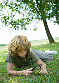 Boy lying in grass, looking through magnifying glass at flower