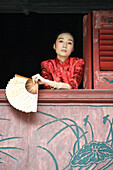 Young woman dressed in traditional Chinese clothing, leaning out of window sill, holding fan