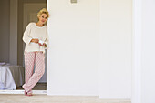 Mature woman leaning against wall in pajamas with cup of coffee