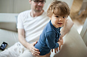 Toddler boy on couch with father, looking at camera
