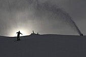 A snow machine spraying snow by a skier on a ski slope