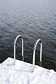 A snow covered dock with a ladder going into rippled water