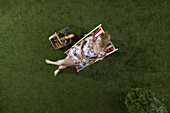 A woman sitting in the sun on a grass in a park with a picnic basket, overhead view