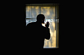 A man peeking outdoors through the blinds of a darkened room