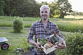 Portrait of smiling mature man carrying crate of freshly harvested vegetables at garden