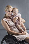 Portrait of happy mother with baby girl in wheelchair against gray wall