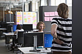 Man standing and stretching by computer desk, colleagues working in background