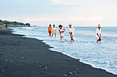 Kinder im seichten Wasser am Strand mit schwarzen Sand, Odalanfest im Tempel Pura Goa Lawah, Padang Bai, Bali, Indonesien