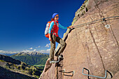 Frau steigt auf Eisenbügel am Drahtseil entlang mit Latemar-Gruppe im Hintergrund, Trans-Lagorai, Lagorai-Höhenweg, Lagorai, Dolomiten, UNESCO Welterbe Dolomiten, Trentino, Italien