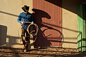 Agriculture - Cowboy with a lasso rope standing against a barn in evening light / Childress, Texas, USA.