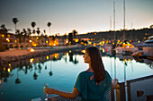 A pretty young girl with a drink in her left hand stands by the marina iron railing.