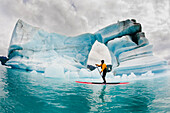 One man on stand up paddle board (SUP) paddles past hole melted in iceberg on Bear Lake in Kenai Fjords National Park, Alaska.
