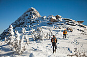 Two climbers wearing backpacks walk a wide snow-covered ridge toward Needle Peak in the Coquihalla Recreation Area of British Columbia, Canada.