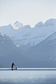 A man stand up paddle boards (SUP) on a calm Lake McDonald at sunrise in Glacier National Park near West Glacier, Montana.