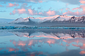 Panoramic view across the calm water of Jokulsarlon glacial lagoon towards snow-capped mountains and icebergs bathed in the last light of a winter's afternoon, at the head of the Breidamerkurjokull Glacier on the edge of the Vatnajokull National Park, Sou