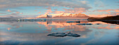 Panoramic view across the calm water of Jokulsarlon glacial lagoon towards snow-capped mountains and icebergs bathed in late afternoon light in winter, at the head of the Breidamerkurjokull Glacier on the edge of the Vatnajokull National Park, South Icela